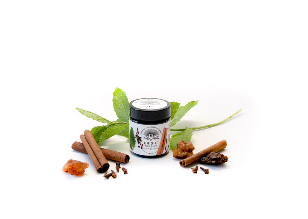 A small jar of Bright tooth polish centered on a white background, surrounded by cinnamon sticks, mint leaves, ginger root, and cloves as flavor ingredients.