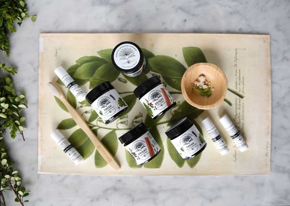 An overhead flat-lay of EAA essentials products arranged on a vintage botanical print with green leaves, a wooden spoon, and a small wooden bowl, set on a light marble surface.