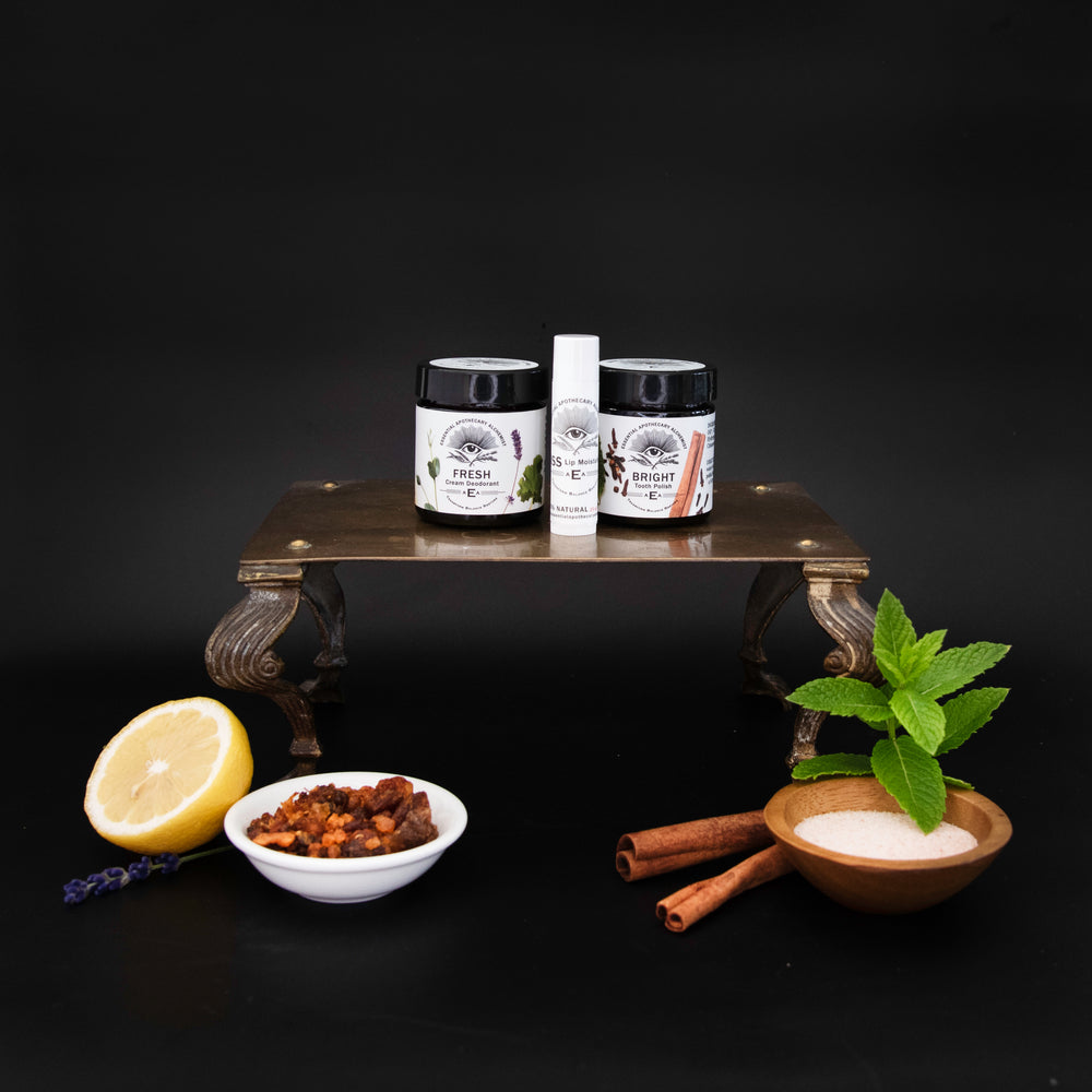 Three essentials products from Essential Apothecary Alchemist displayed on a dark wooden stand against a black background, with lemon, dried fruit, cinnamon sticks, mint, and a bowl of white scrub arranged around them as ingredients.